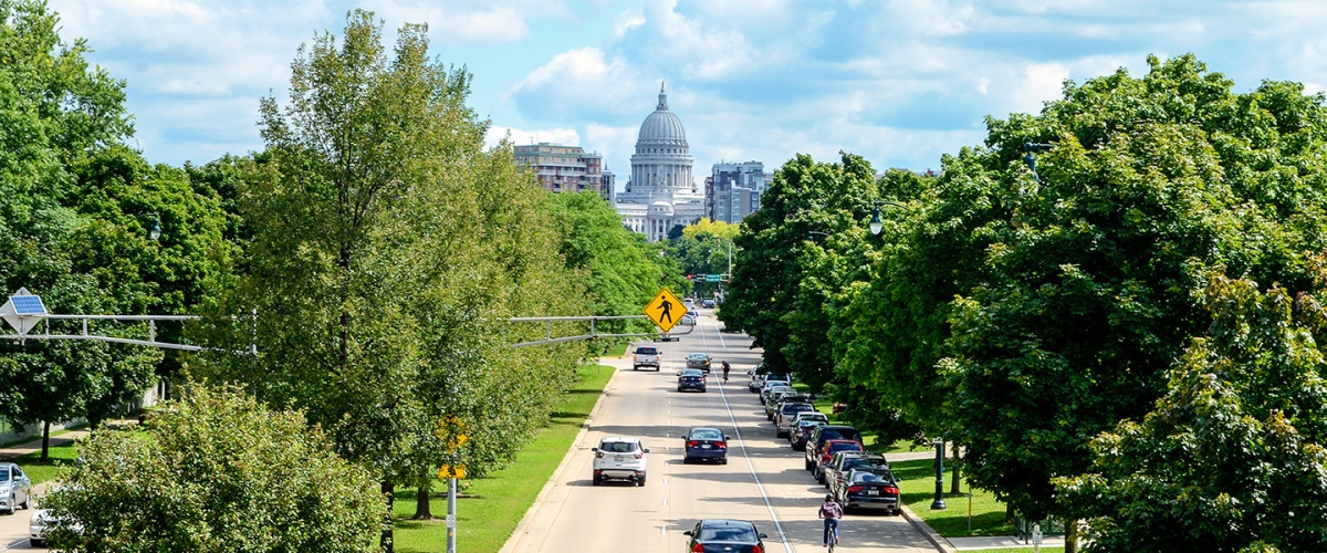 City street leading towards the Wisconsin Capitol Building in the distance, with cars driving and parked on the street, flanked by full, green trees.