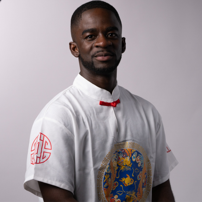 A black ,am wearing short cropped hair and beard looks into the camera. He is wearing a white shirt with a red satin tie closure at the next and a decorative motif on the chest and shoulder.,