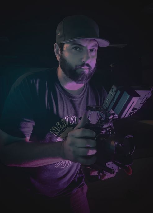 A man with a baseball cap with a beard and a grey t-shirt holds a camera