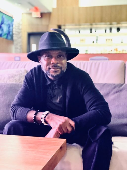 Man with a black wide-brimmed hat and black clothes sits on a couch in front of a coffee table.