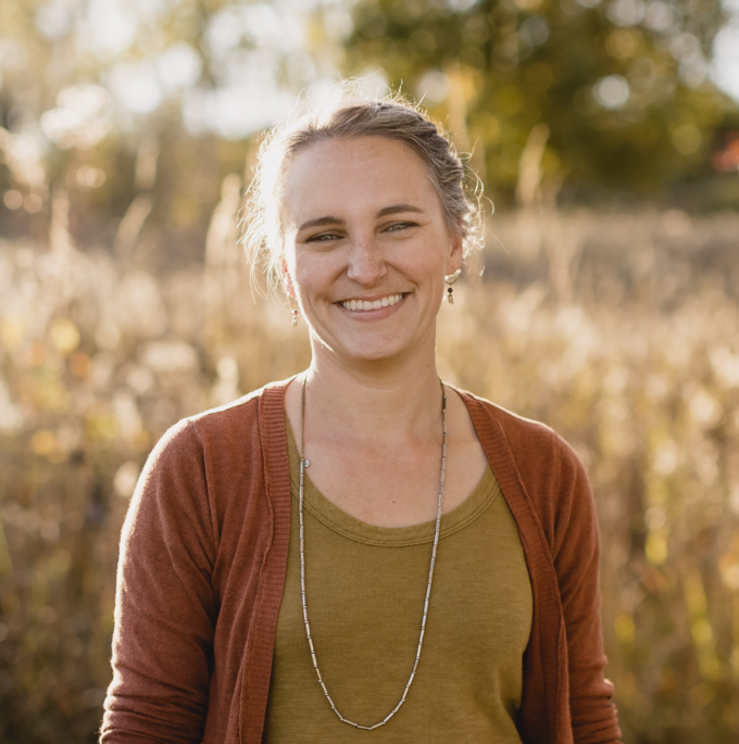 Photo of a person standing in a field with a yelow shirt, orange sweater, and a silver necklace Her hair is tied back and she is smiling at the camera. 