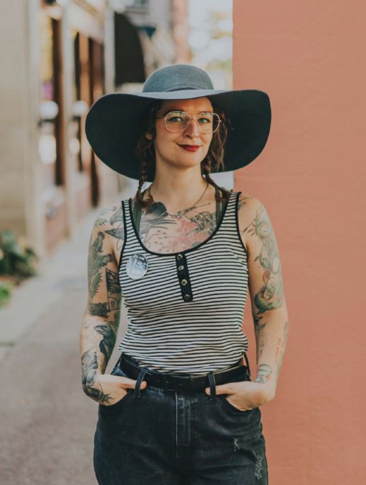 Photo of a person with a black broad rimmed hat, wire frame glasses, and red lipstick standing with their hands in the pockets of their black jeans.