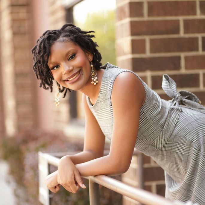 A woman with a grey dress leans against a railing and smiles at the camera