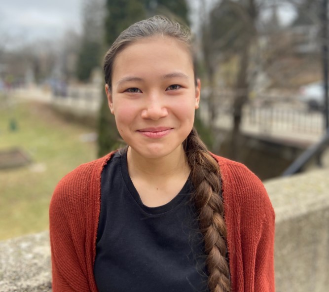 2023 Madison Youth Poet Luareate Madeleine Bohn is pictured wearing an orange sweater over a black shirt with her brown hair in a braid down her shoulder.
