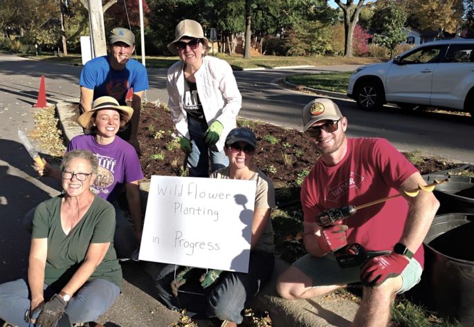 Six people crowd together and smile at the camera in front of a planting bed. A person in the middle holds a sign that reads pollinator planting in progress.