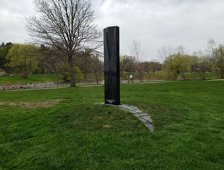 A shiny black structure placed on a half moon stone path in a green field.