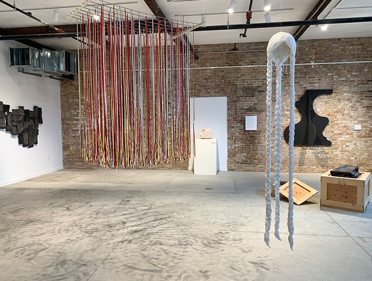 Industrial space with abstract art hanging on the wall in the background, the foreground has two intricate pieces with colorful string and a long braided hairpiece.
