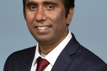 Alder Badri Lankella, male of Indian descent, is wearing a dark gray suit, with a red tie, white collared shirt, with short black hair.