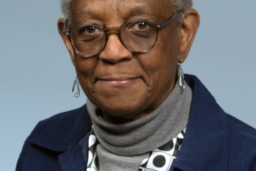 Alder Joann Pritchett, of African American descent, is wearing a dark blue denim jacket, white and gray polkadot sweater and a gray turtleneck under the jacket.