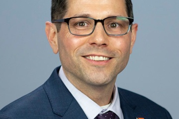 Alder John Duncan, a Caucasian male, is wearing a blue blazer, with a dark red tie, short black hair and dark rimmed glasses.