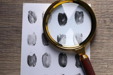 A sheet of white paper with fingerprints lies on a table with a magnifying glass on top of it. 
