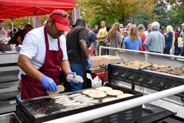 Man in a red apron grilling food outdoors at an event.
