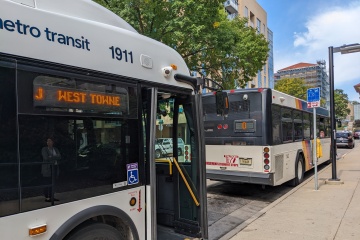 Two buses waiting at a bus stop with one displays ‘J West Towne’ on marquee.
