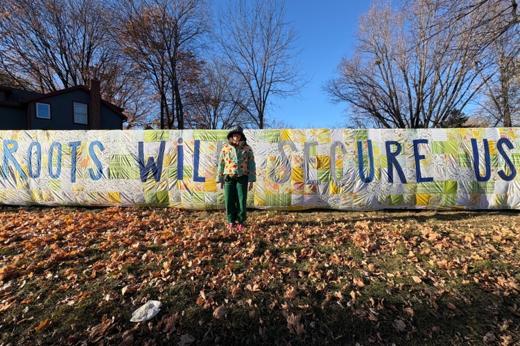 A person standing in front of a large quilt with the phrase "Roots Will Secure Us"