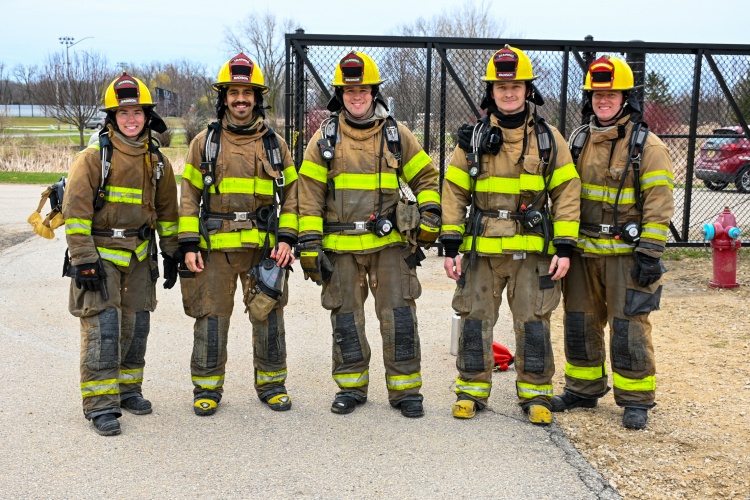 Five firefighter recruits stand in turnout gear and smile to the camera