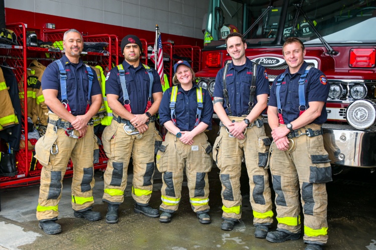 Firefighters stand in a row in front of a fire truck and gear locker