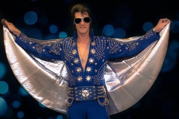 A man with dark hair and sunglasses wearing a shimmery blue outfit and a white cape.