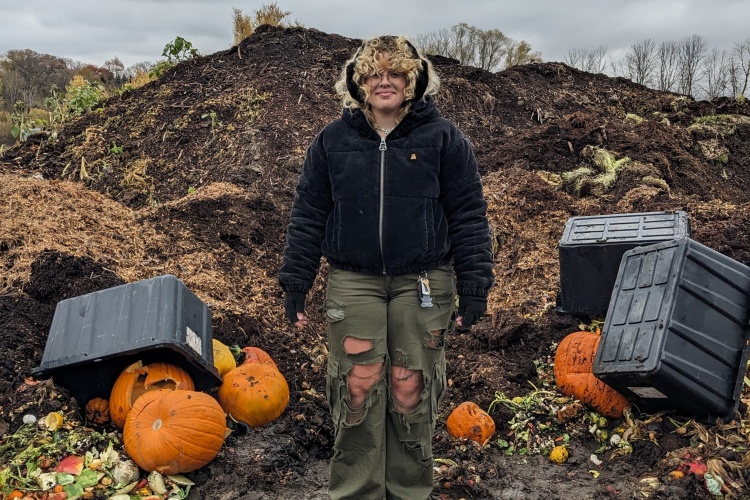 Intern with ripped jeans standing in front of a very muddy compost pile. There are overturned plastic totes near her. 