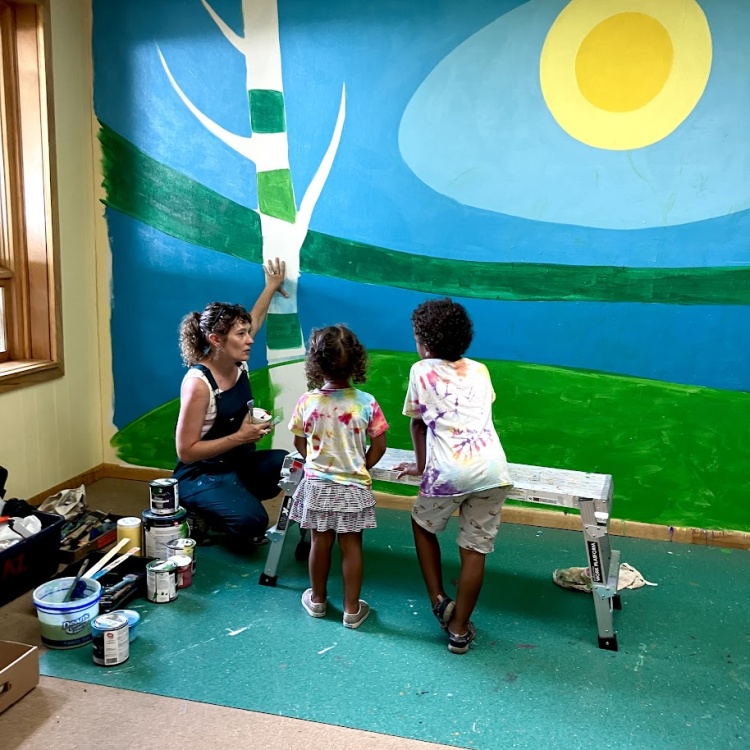An adult showing two children how to paint a mural of a tree and a sun.