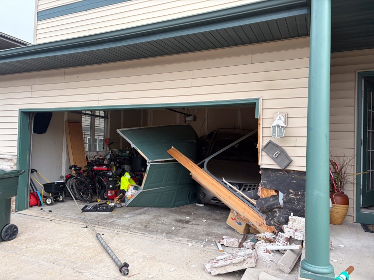 A vehicle rests in the garage with the garage door leaning against it and other damaged property nearby