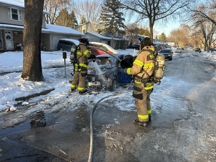 Firefighters spray water and foam on the front hood of a blue sedan