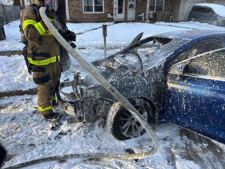 Up-close view of a firefighter applying water and foam to the engine compartment of a blue sedan