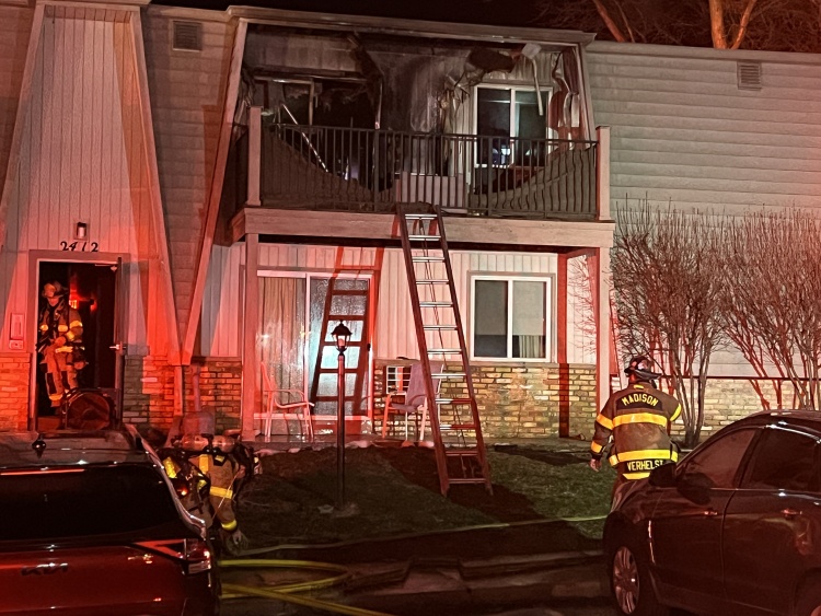 Firefighters stand in front of building with a ladder leaning against a balcony that has been damaged by fire