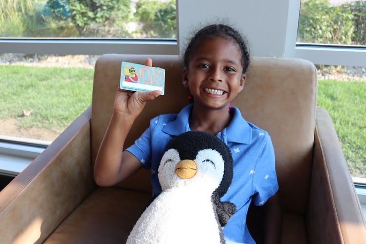 Un niño se sienta con un pingüino de peluche en su regazo, sonriendo y sosteniendo una tarjeta de edición especial de la biblioteca.