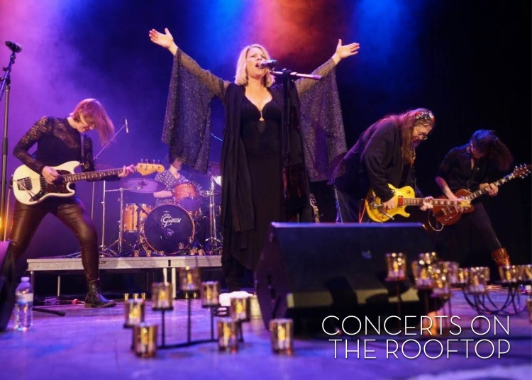 5 -woman band all dressed in black, 1 on drums, 3 on guitar, 1 lead singer standing centerstage with arms raised in the air as she sings