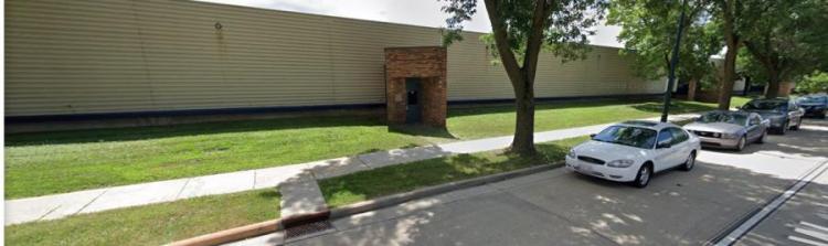 A photo of a tan colored metal wall and brick door enclosure with green grass in front of the structure and a tree in the avenue between the sidewalk and the road