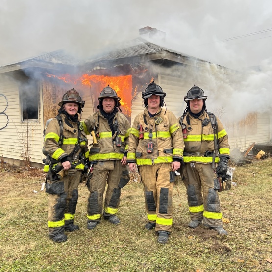 Four firefighters stand in front of a burning house