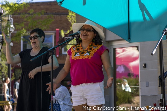 A latina woman wearing a hat stands at a mic telling a story during a story slam hosted by Madison Public Library as part of Library Takeover.