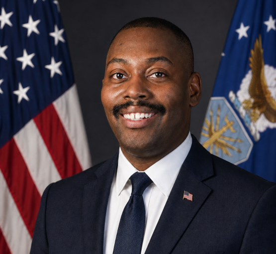 A formal portrait of Gregory McClain, who is wearing a dark suit and is in front of two American flags 