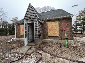 A brick house with boarded-up windows and large letter "A" spray painted on the front