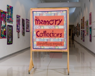 Sign reading "Memory Collectors Storytelling Project" sits in a gallery hallways featuring quilts on display. 