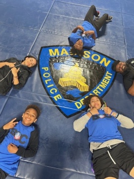 Four middle school students and an officer smile around the Madison Police Department logo at a summer camp hosted by our department.