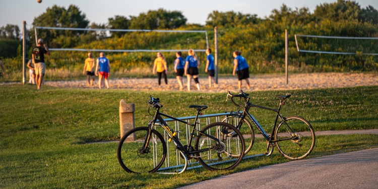 bicycles resting in a bike rack as adults play volleyball on the sand court. 