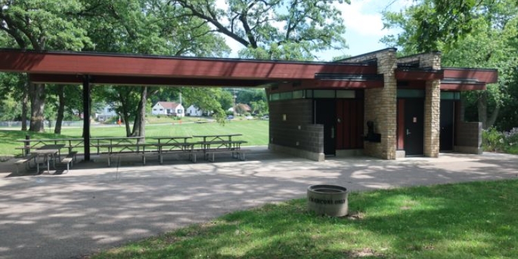 Shelter with restrooms and picnic tables surrounded by lawn and trees.
