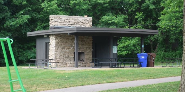 Shelter with picnic tables. There is a lawn with a sidewalk and there are trees behind the shelter.