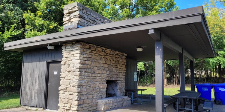 Shelter with picnic tables. There is a lawn with a sidewalk and there are trees behind the shelter.