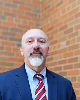 Bob Soldner, Caucasian male with gray beard, Blue blazer, light blue shirt, with red and white striped tie, standing in front of a brick wall