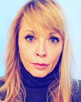 Michelle Drea, blonde shoulder length hair, wearing a black turtle neck, wiith light blue background