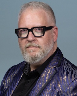 Alder Mayer is wearing a purple blazer with black thick rimmed glasses.