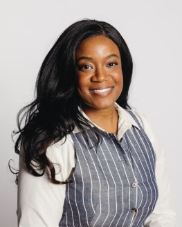 Alder Devine Nzegwu is young Black woman with long Black hair. She is wearing a white collar shirt with a gray vest.
