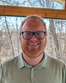 Alder Elect Noah Lieberman is wearing dark rimmed glasses, a green shirt and is smiling while standing in front of a window with trees in the background.