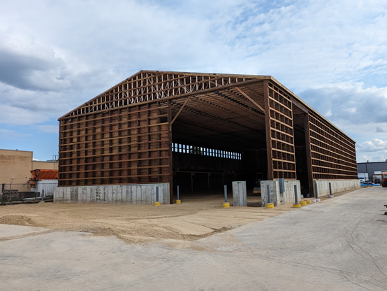 This is another snapshot of the under construction salt storage facility. The interior is closer to finished as the floor and walls are nearing completion. Markings on the wall note how full salt can be piled.   The structure has a concrete base and wooden sides and a roof.  It has a wooden crosshatch design across the front and side and two large openings for trucks.